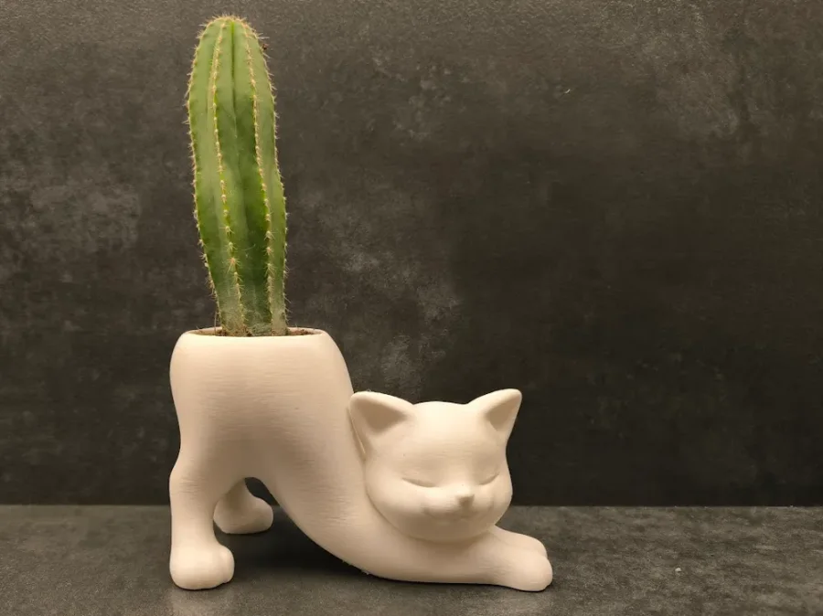 Hvit 3D-printet blomsterpotte formet som en avslappet katt med lukket øyne og integrert potte i ryggen, vist med en høy grønn kaktus.