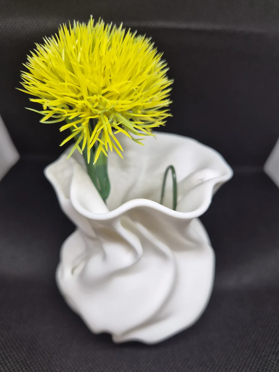 Liten lys vase med avrundet kropp, organiske folder og bølgende åpning, vist med en gul blomst mot mørk bakgrunn.