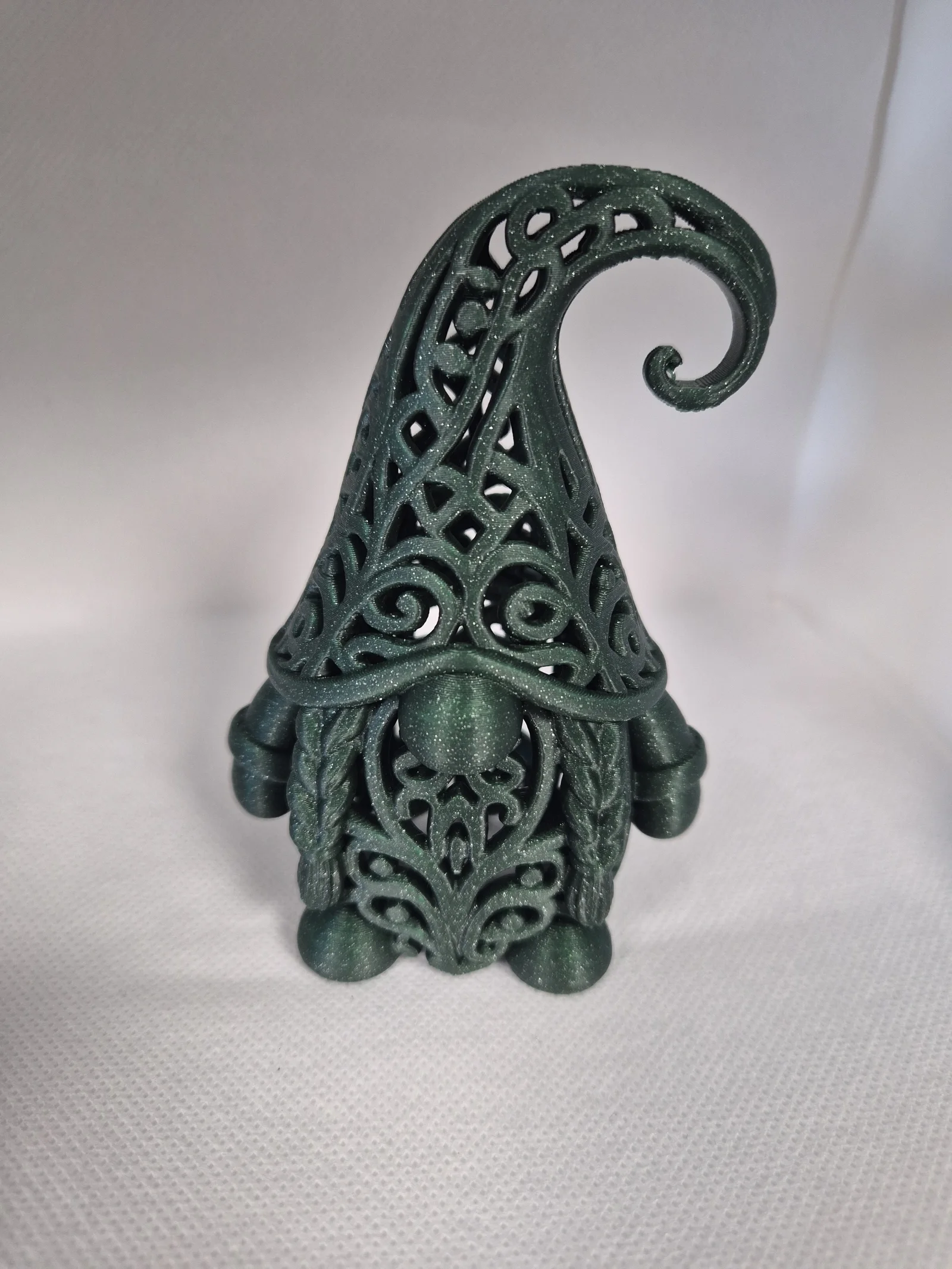 Mørkegrønn glitrende julefigur formet som en nisse med åpent ornamentmønster, rund nese, skjegg og høy spiralformet lue.