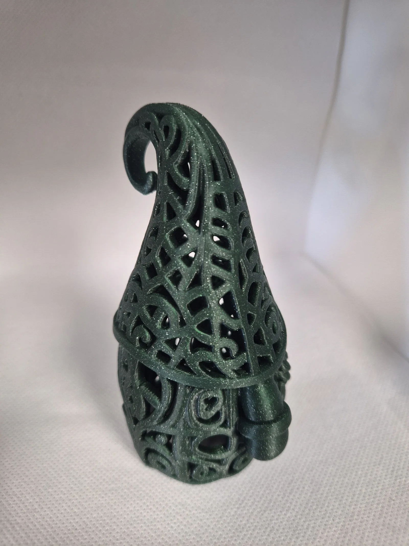 Mørkegrønn glitrende julefigur formet som en nisse med åpent ornamentmønster, rund nese, skjegg og høy spiralformet lue.
