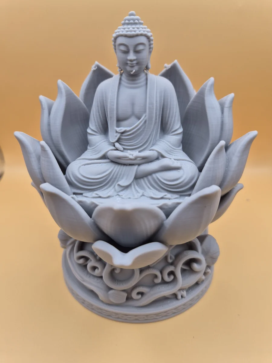 Lys grå Buddhafigur i meditativ sittestilling, omgitt av lotusblader og plassert på en ornamentert rund sokkel mot varm bakgrunn.