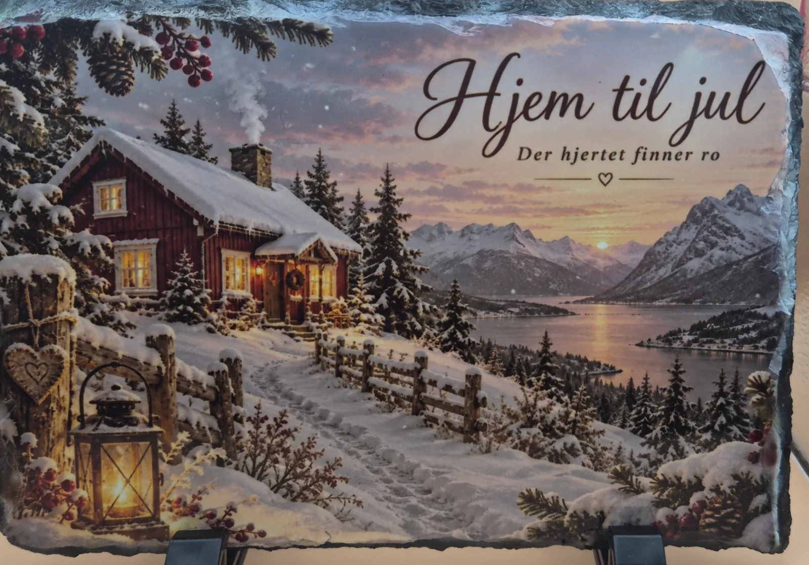 Rektangulær skiferplate med ujevn mørk kant, plassert i et svart stativ. Motivet viser en snødekt rød hytte ved fjell og fjord, en tent lykt og teksten «Hjem til jul – Der hjertet finner ro».