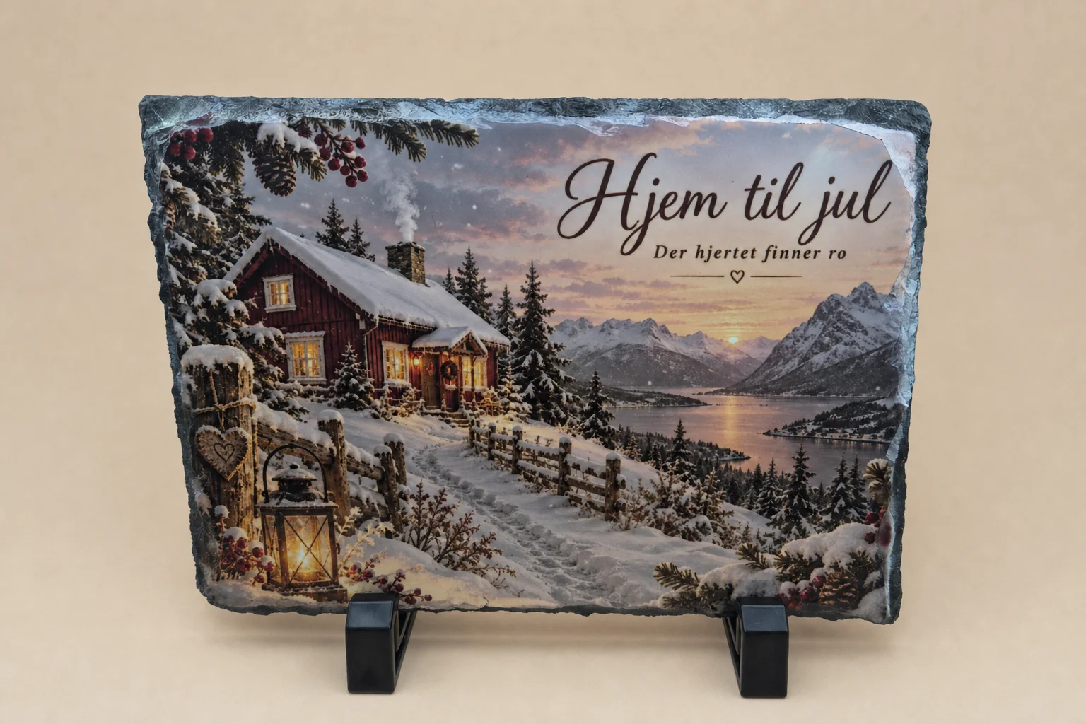 Rektangulær skiferplate med ujevn mørk kant, plassert i et svart stativ. Motivet viser en snødekt rød hytte ved fjell og fjord, en tent lykt og teksten «Hjem til jul – Der hjertet finner ro».