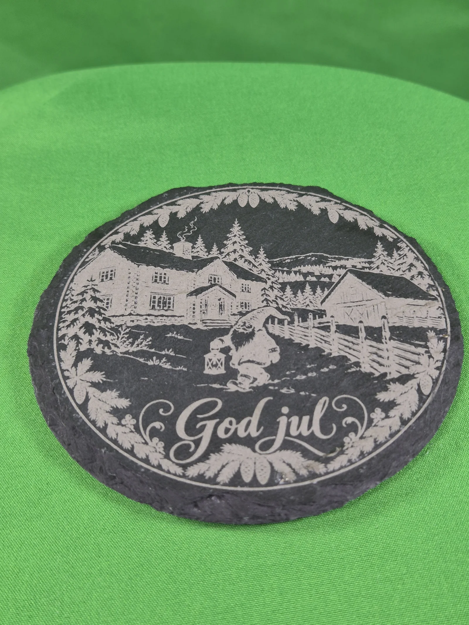 Rund, mørk drikkebrikke i skiferstein på grønt tekstil, lasergravert med vinterlig gårdstun, nisse med lykt, granbar og teksten «God jul».