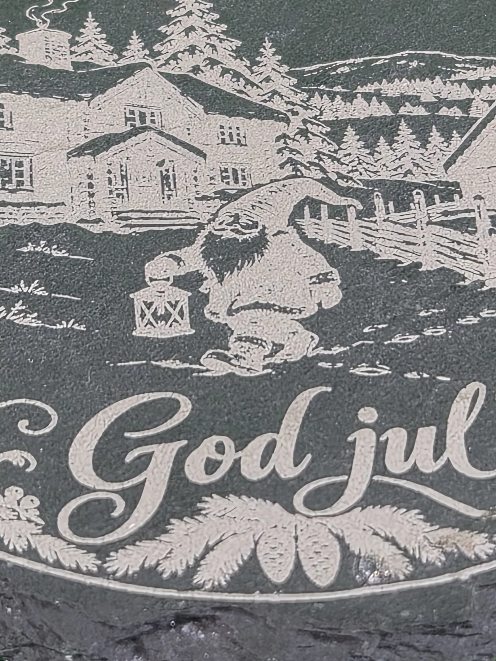 Rund, mørk drikkebrikke i skiferstein på grønt tekstil, lasergravert med vinterlig gårdstun, nisse med lykt, granbar og teksten «God jul».