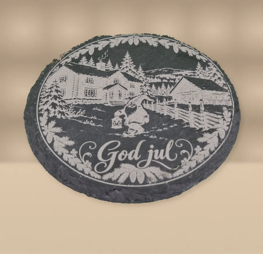 Originalfoto av en rund, mørk drikkebrikke i skiferstein med lasergravert vintermotiv og teksten «God jul», på en lun sandbeige studioflate.